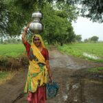 Indian woman in colorful sari carries metal pots through rural field, showcasing traditional lifestyle.