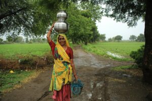 Indian woman in colorful sari carries metal pots through rural field, showcasing traditional lifestyle.