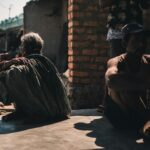 A group of adults sitting outdoors in a village setting, casting strong shadows on a sunny day.