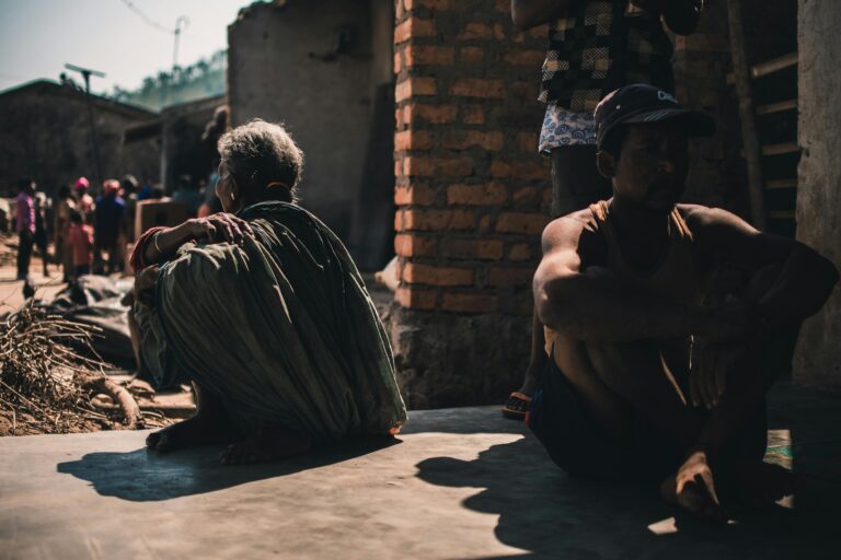 A group of adults sitting outdoors in a village setting, casting strong shadows on a sunny day.
