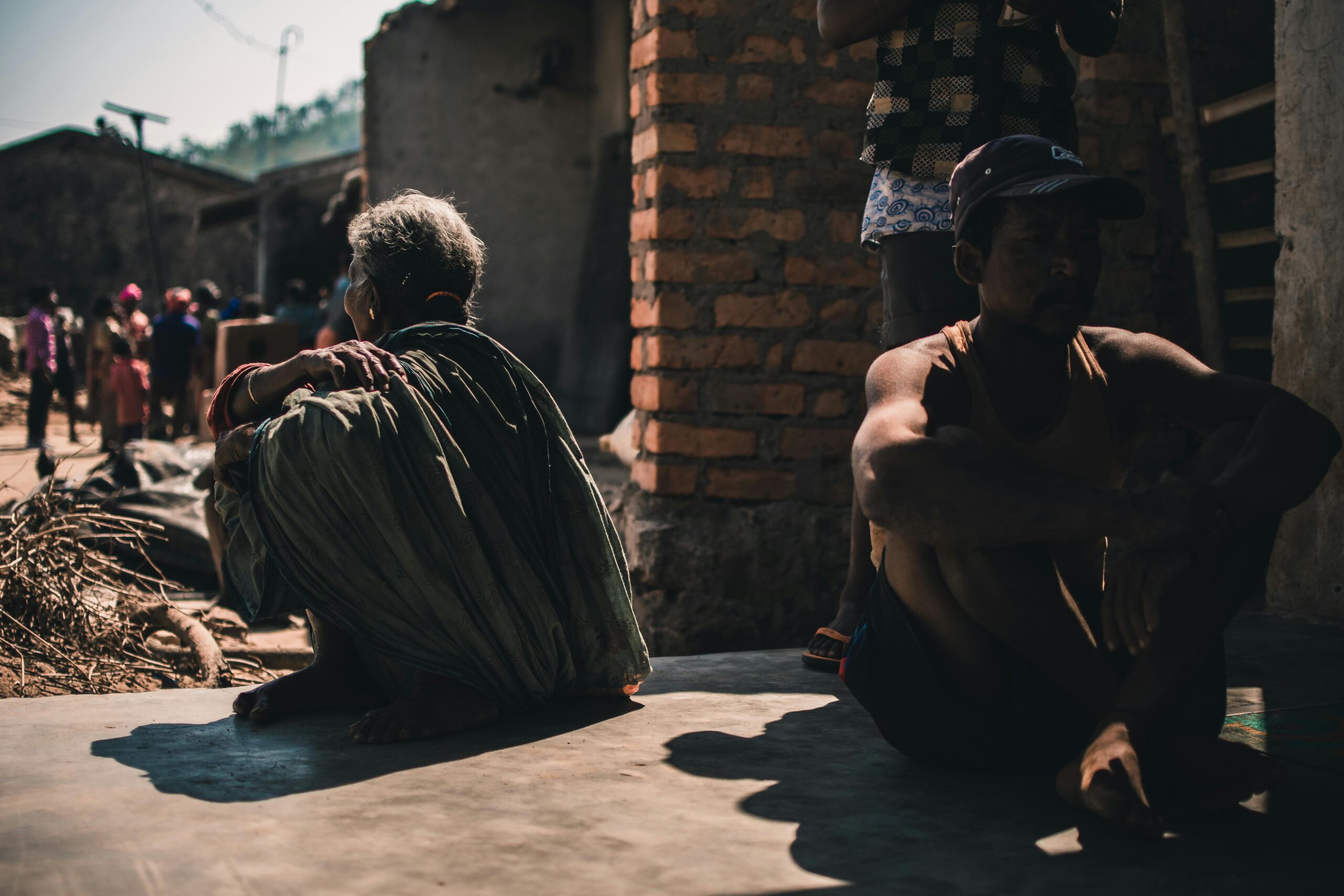 A group of adults sitting outdoors in a village setting, casting strong shadows on a sunny day.