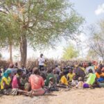 A diverse group of adults and children gather under a tree for a community meeting outdoors.